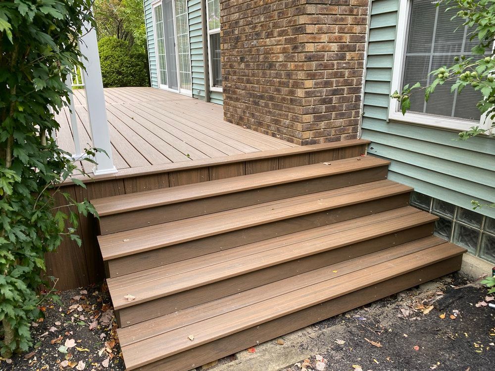 Brown deck and stairs leading up to a house with a brick chimney. Green siding and windows.