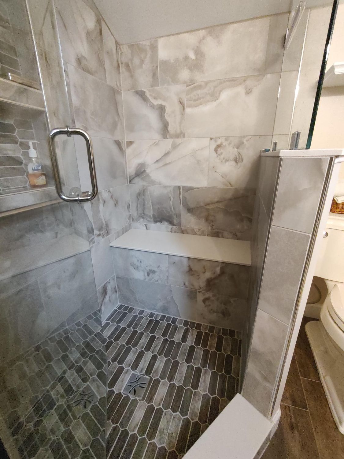 Newly tiled shower with gray marble-look walls, built-in seat, glass door, and mosaic floor.