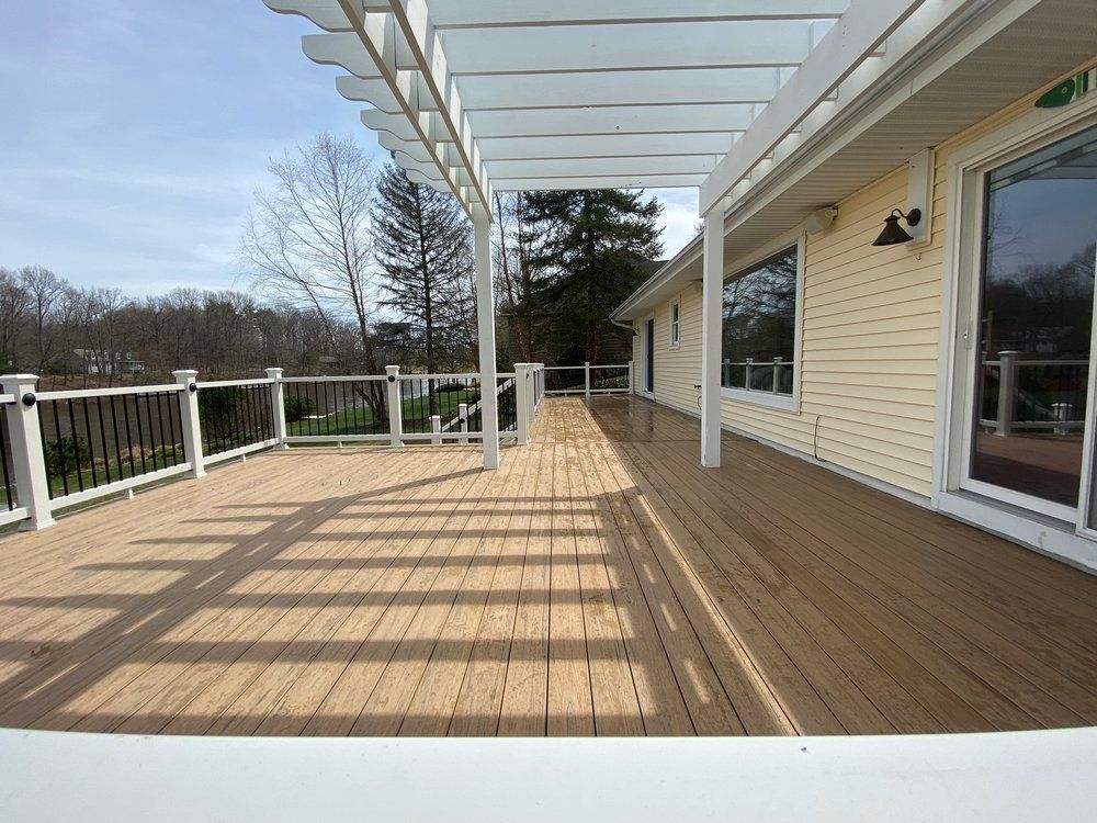 A wooden deck with a pergola, beside a light-yellow house, on a sunny day.
