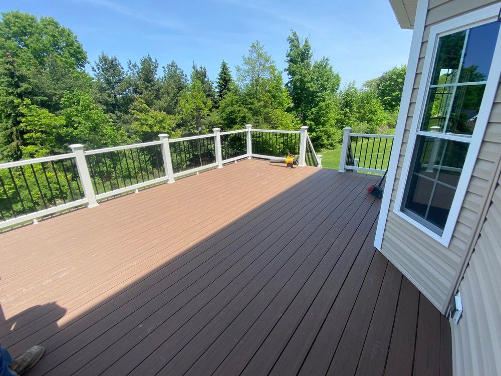Brown composite deck with black railing and trees under a blue sky. A window is on the right.