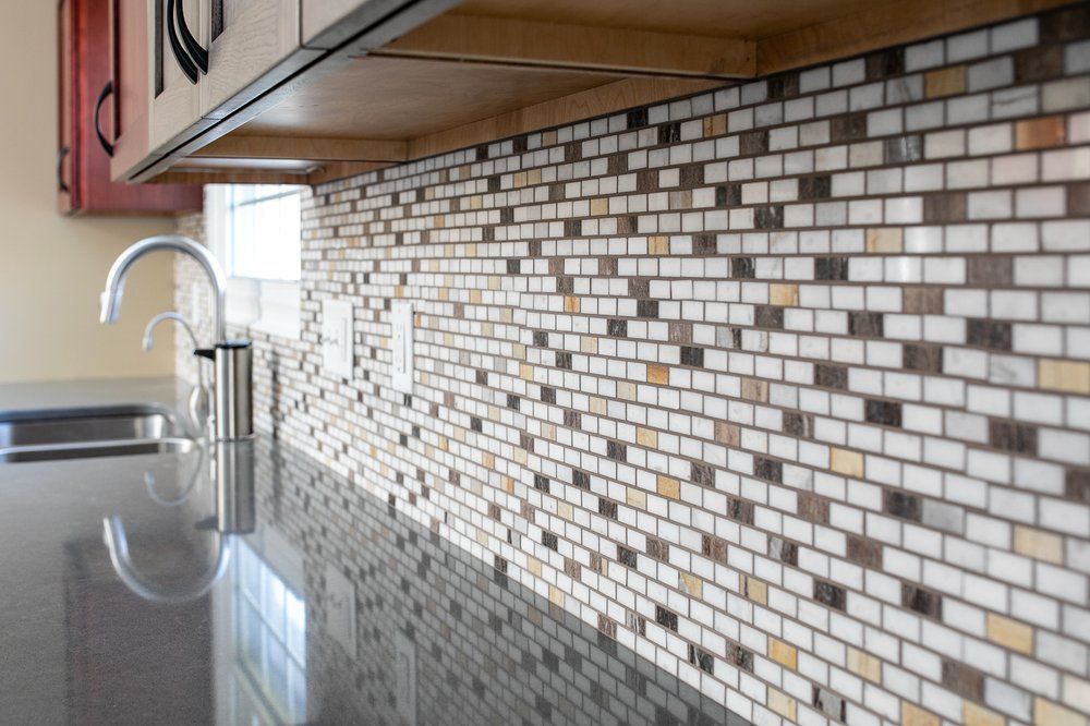 Kitchen backsplash of varied beige, brown, and white brick-shaped tiles; dark countertop, silver faucet.