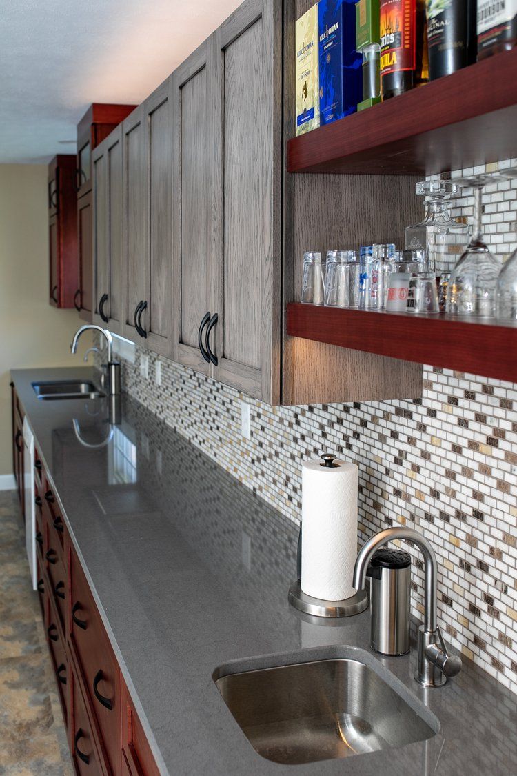 Long bar with dark gray countertop, two sinks, mosaic backsplash, cabinets, and shelves holding glassware and bottles.