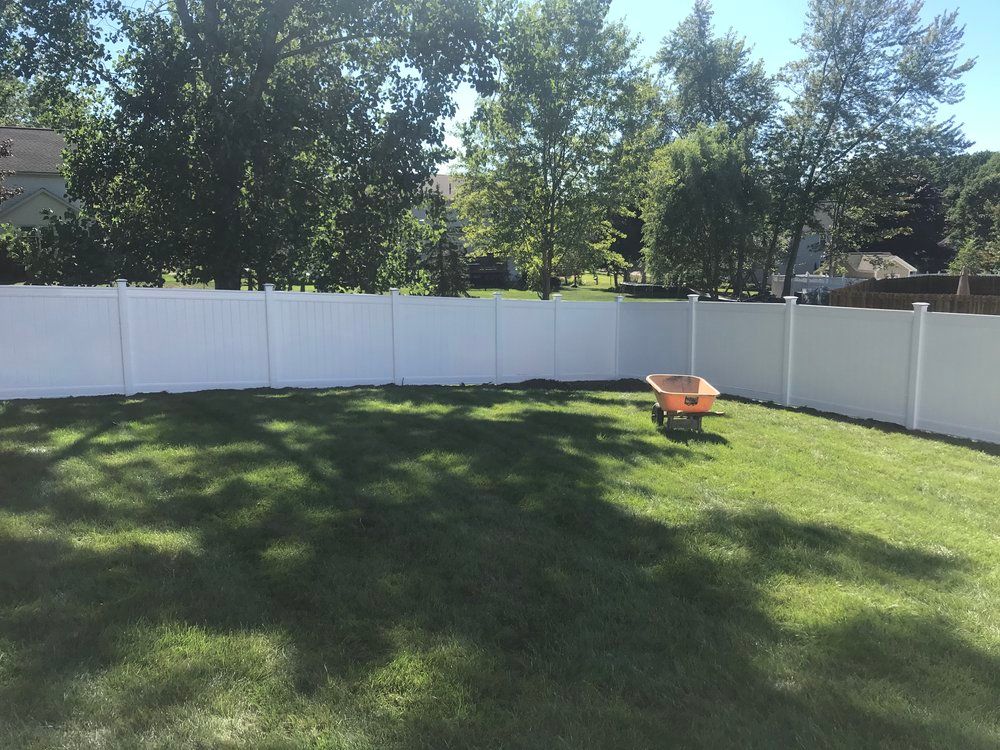 White vinyl fence encloses a grassy backyard with a wheelbarrow. Trees and blue sky in the background.