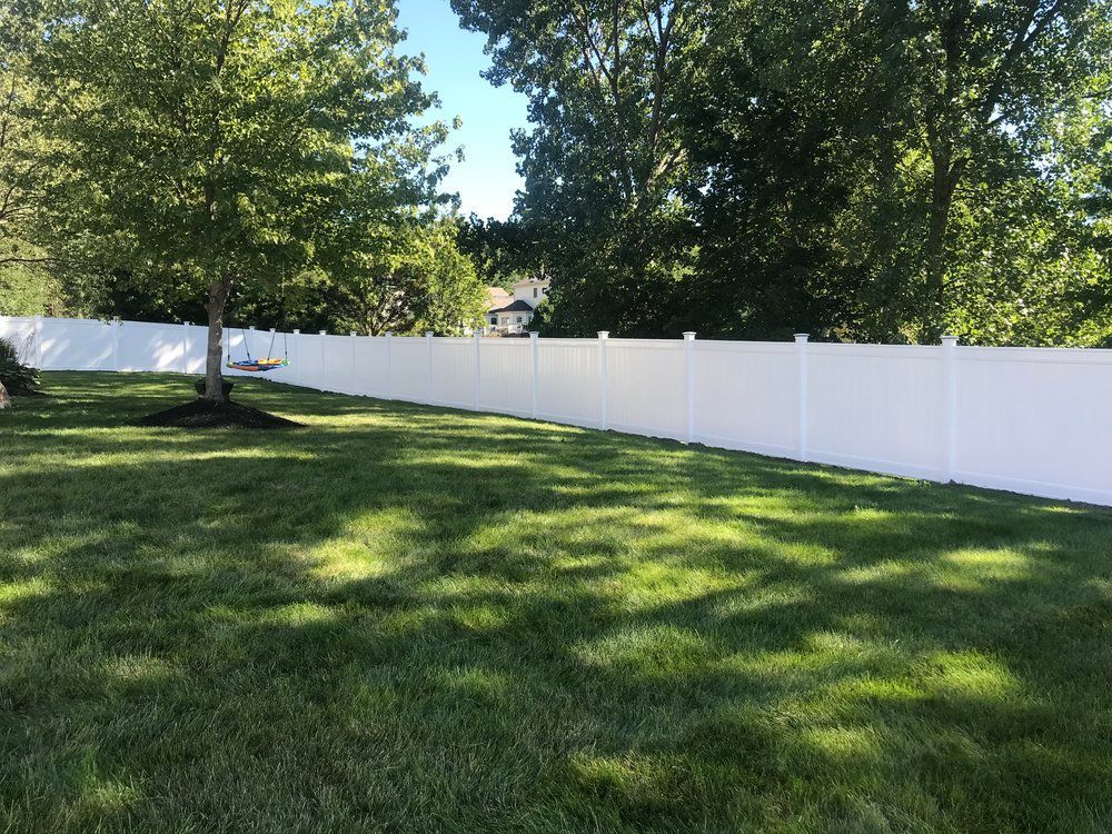 A white vinyl fence bordering a grassy lawn, trees, and blue sky.