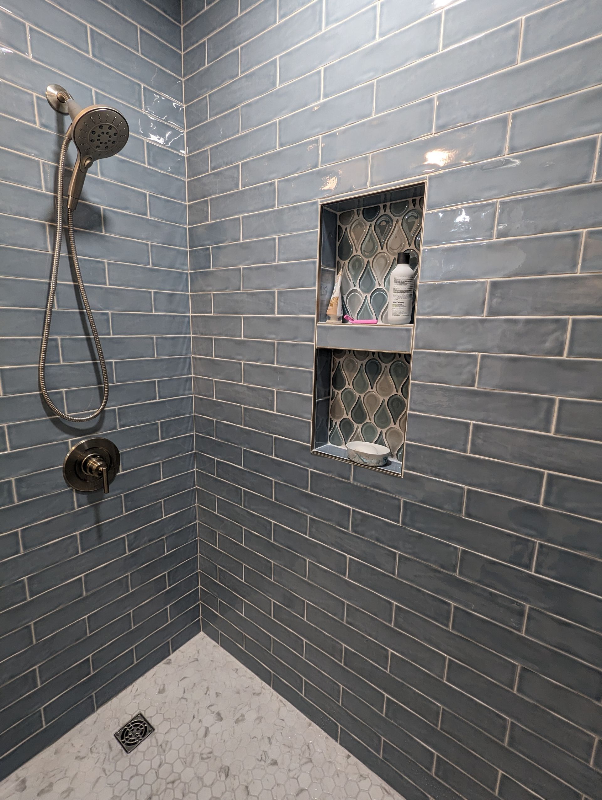 Blue tiled shower with a niche, holding toiletries, and silver fixtures.