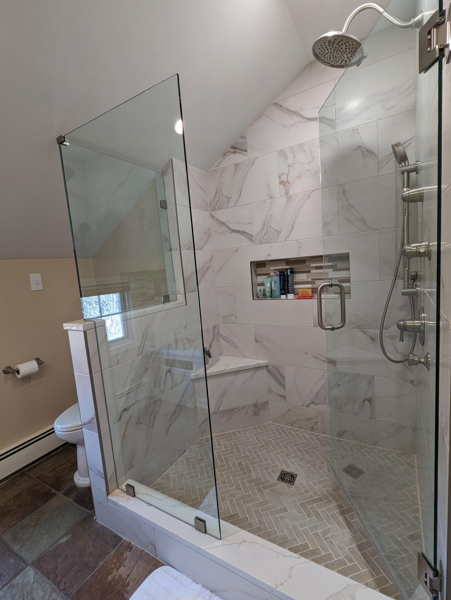 Shower with glass doors and white marble tile. Has a built-in bench and a recessed shelf.