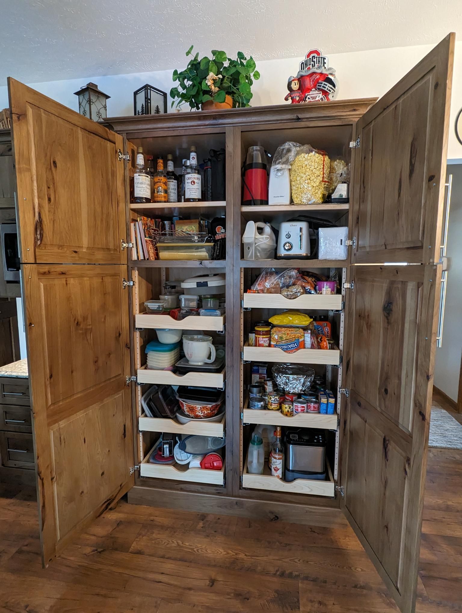 Wooden pantry cabinet with open doors, filled with various food items and kitchen supplies.