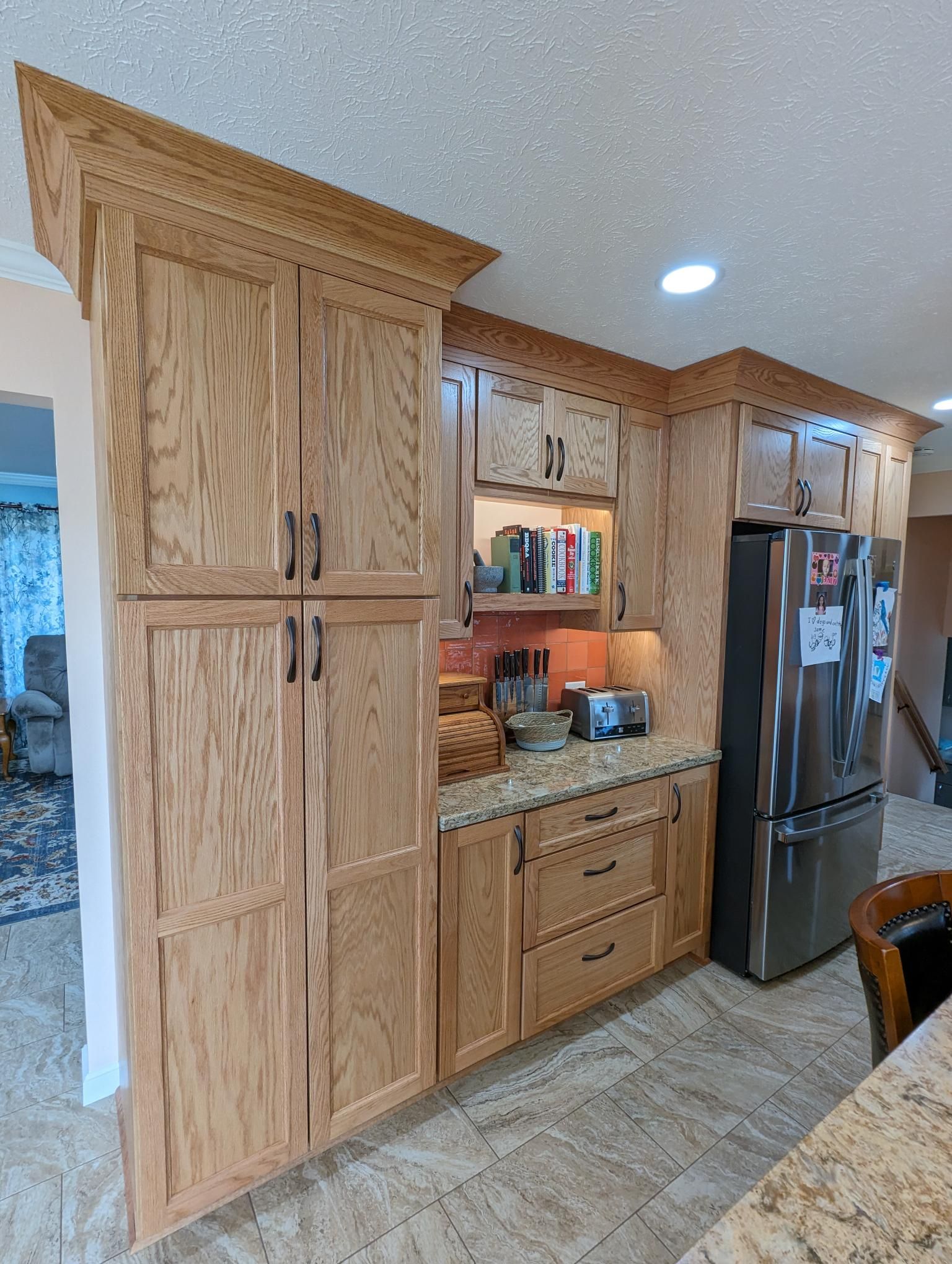 Wooden kitchen cabinets and a stainless steel refrigerator in a kitchen with beige countertops and tile flooring.