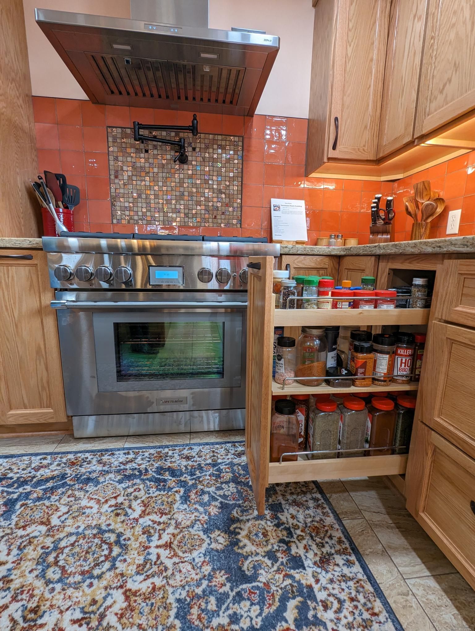 Kitchen with stainless steel oven, spice rack, and orange backsplash; rug in foreground.