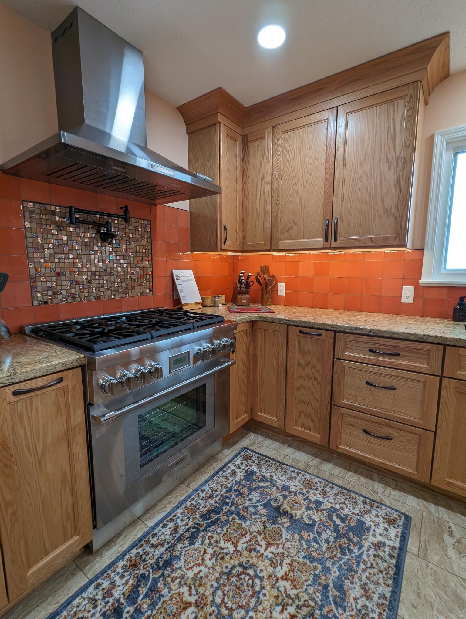 Kitchen with stainless steel range, wooden cabinets, orange backsplash, and blue rug.