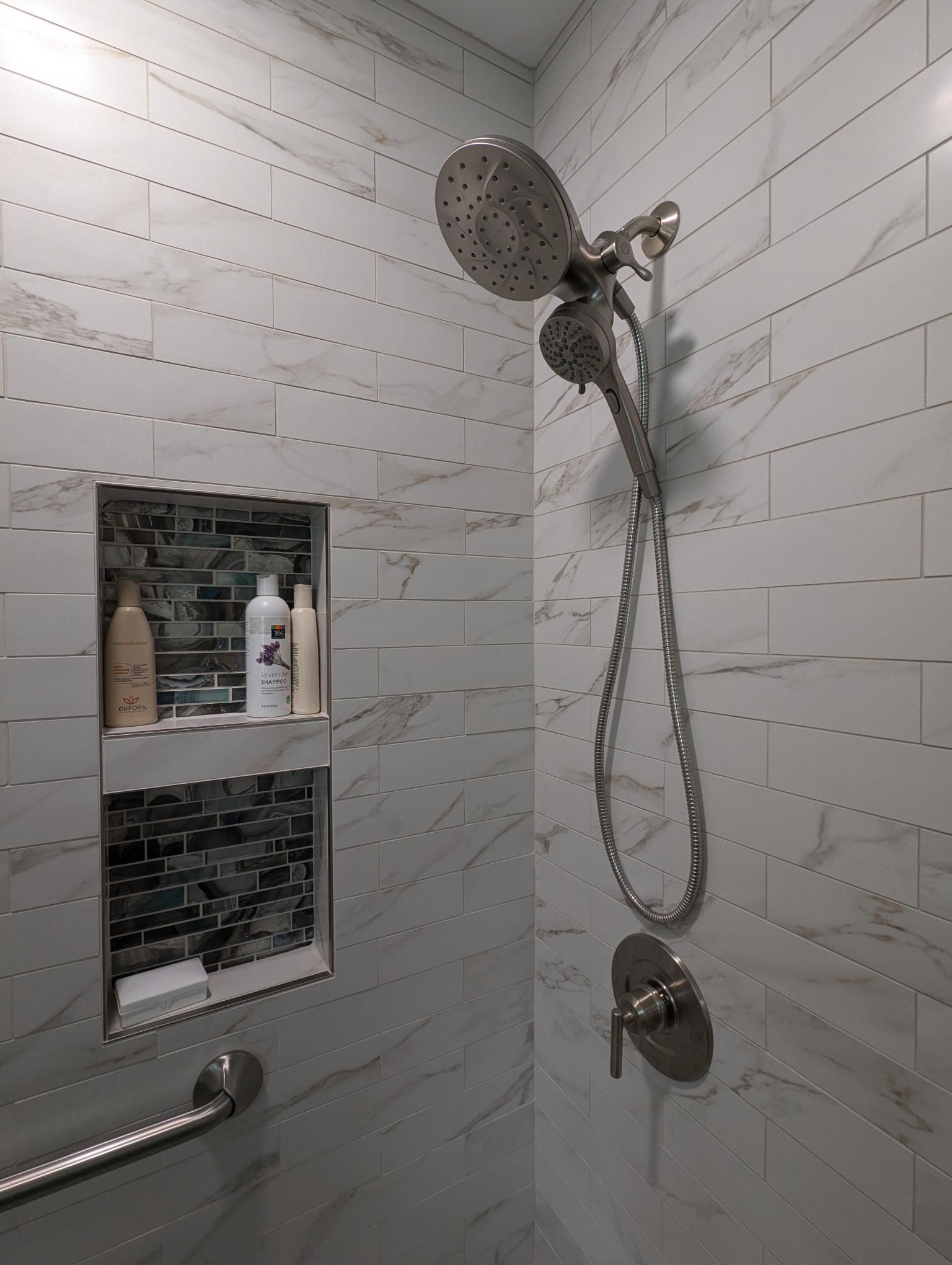Tiled shower with handheld and overhead showerheads, recessed shelf with toiletries, and a grab bar.