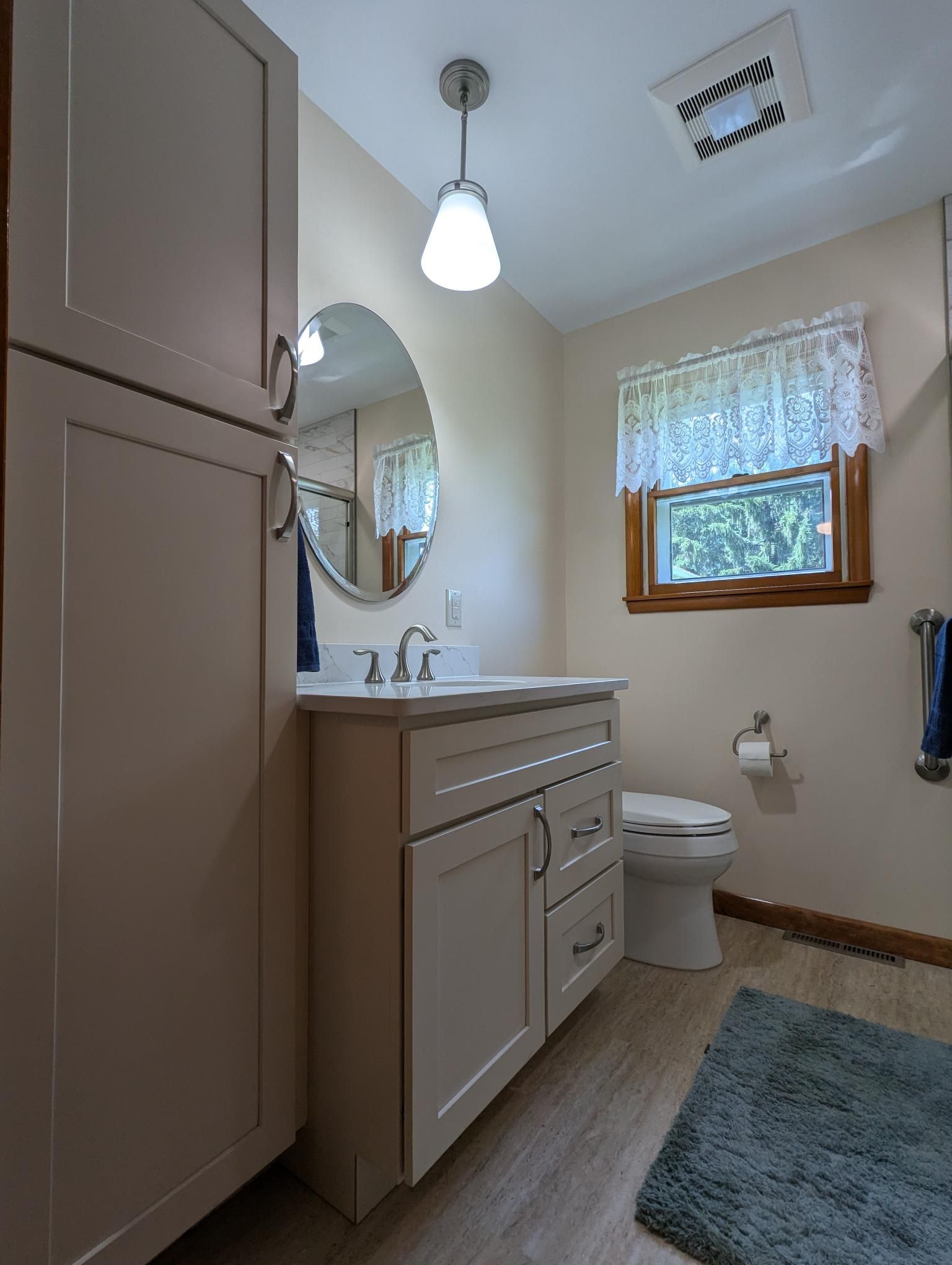 Bathroom with light-colored cabinets, vanity, and toilet. Oval mirror, window with lace curtain, and blue bath mat.