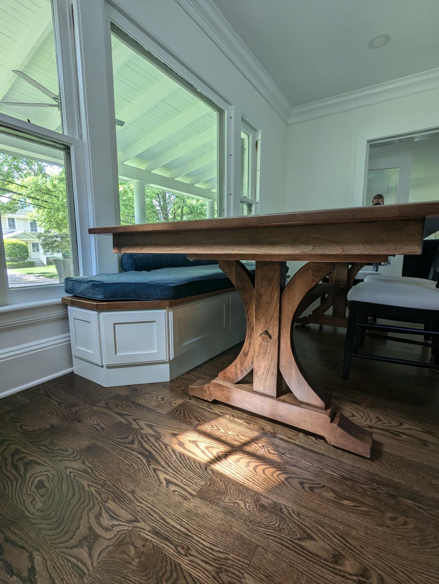 Wooden dining table in a sunlit room with built-in bench seating and large windows.