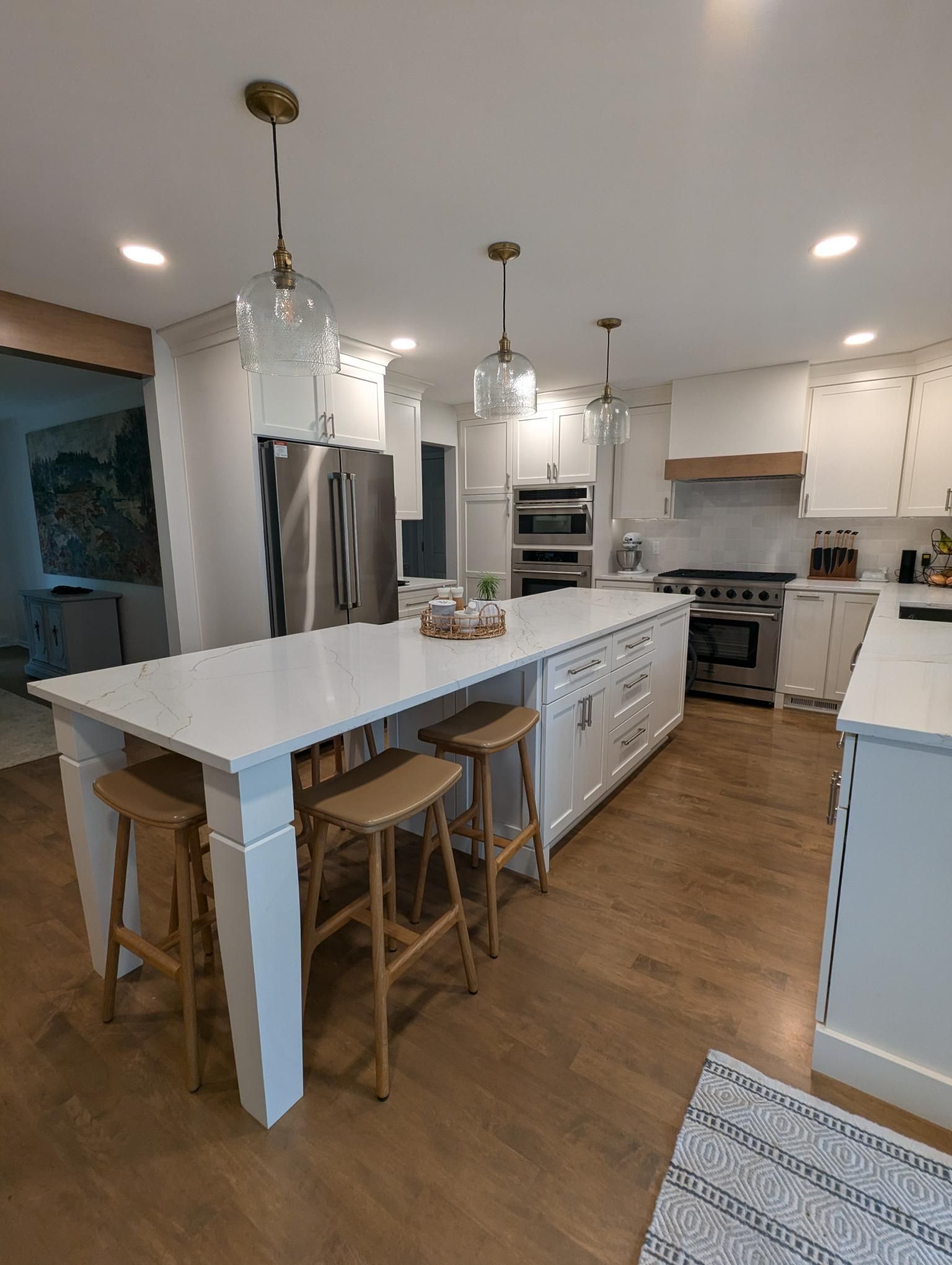Modern white kitchen with island, pendant lights, stainless steel appliances, and wooden stools.