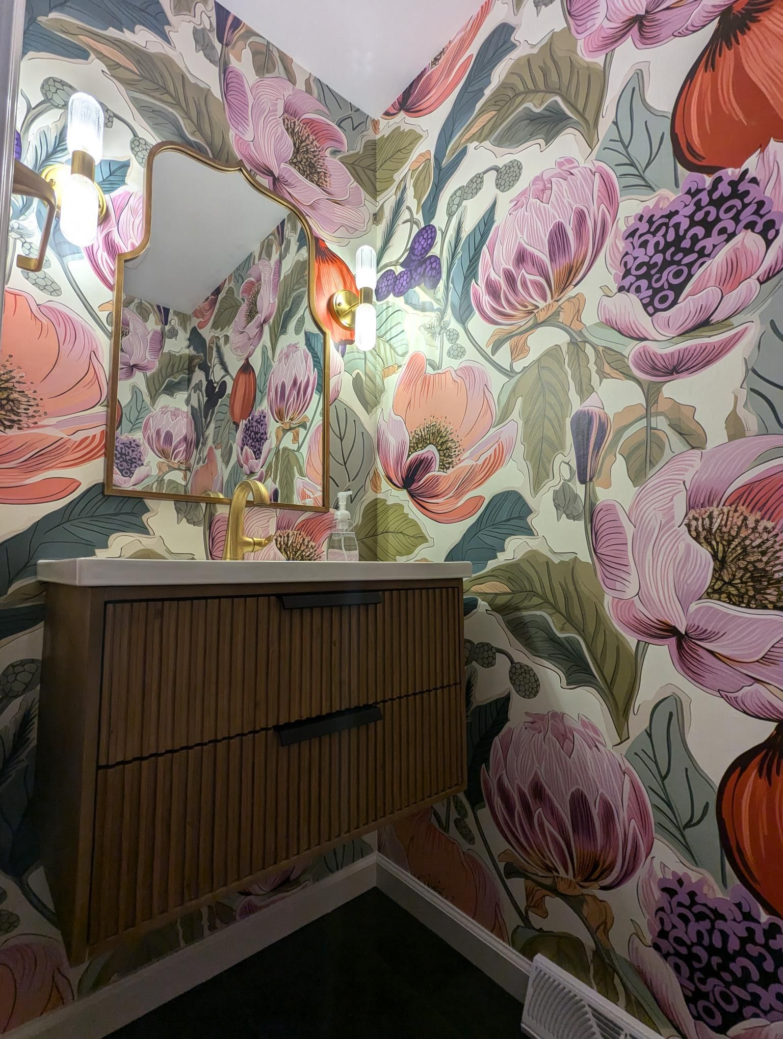 Small bathroom with floral wallpaper, wood vanity, and gold-framed mirror.