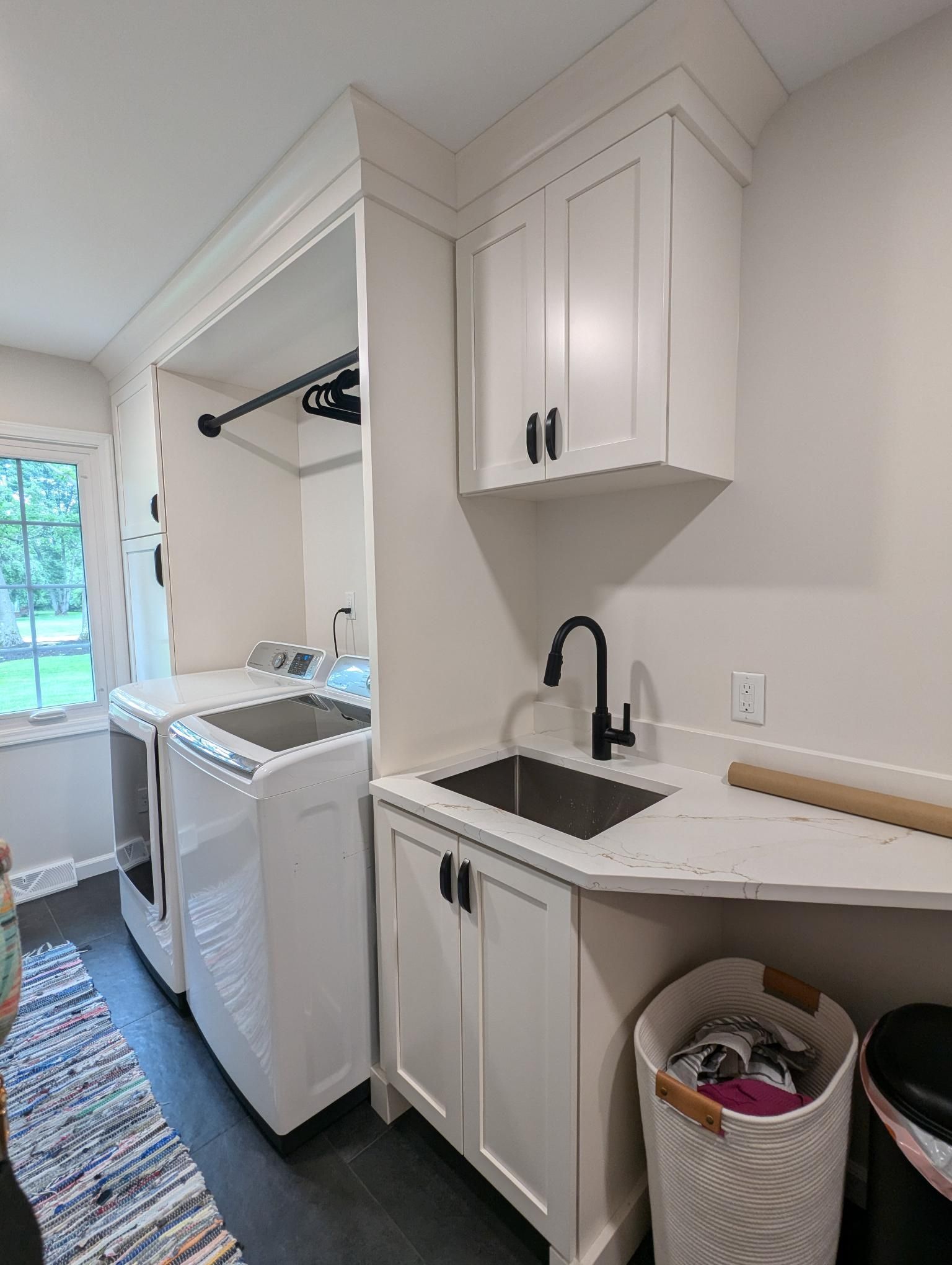 Laundry room with white cabinets, black faucet, sink, washer, dryer, and clothes hamper.