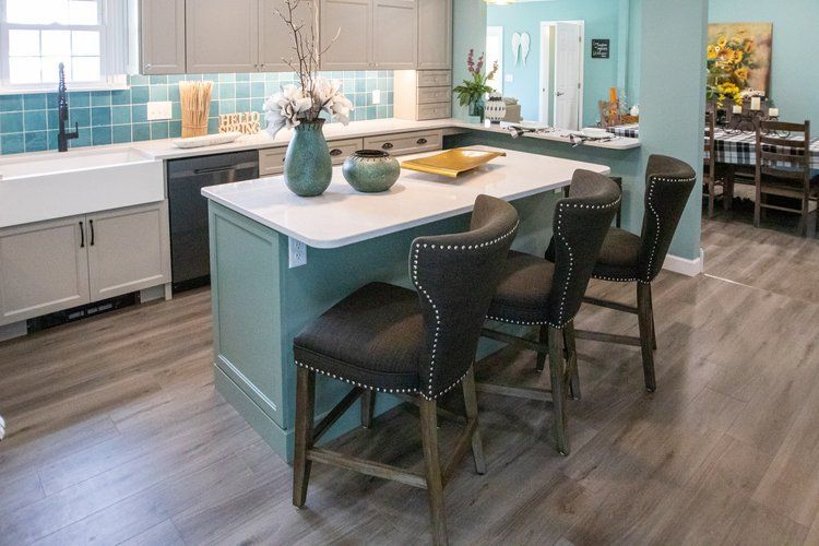 Kitchen with teal island, white countertop, gray cabinets, teal backsplash, wooden floors, and three bar stools.