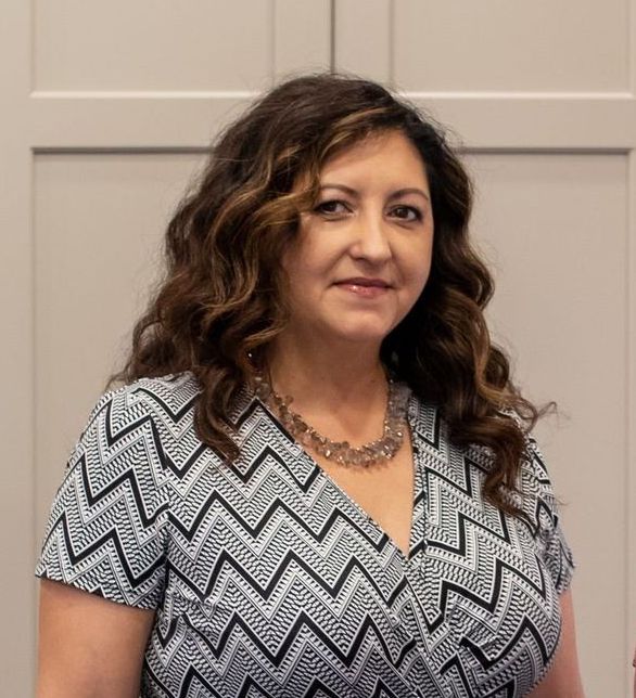 Woman with curly brown hair wearing a patterned dress, smiles in front of a light wall.