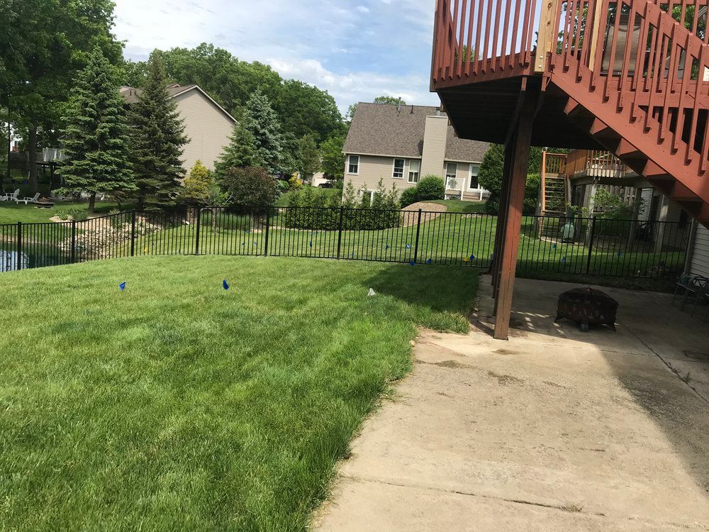 Backyard with green grass, black fence, and a deck. Houses and trees in the background under a blue sky.