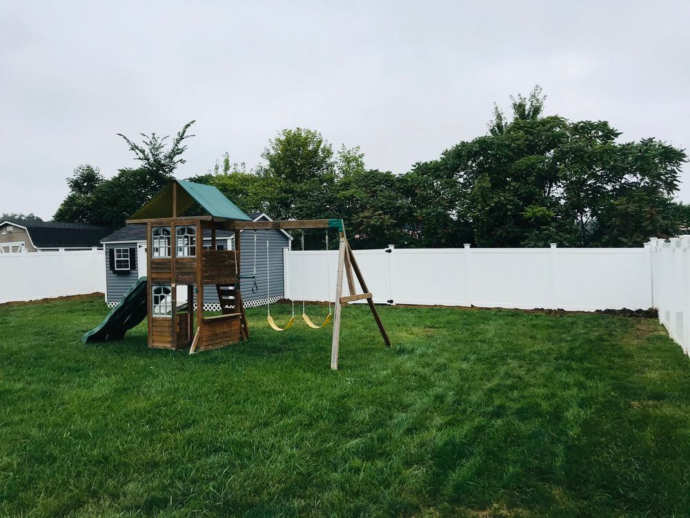 Backyard with a wooden playset, swings, and white fence. Green grass and trees under an overcast sky.