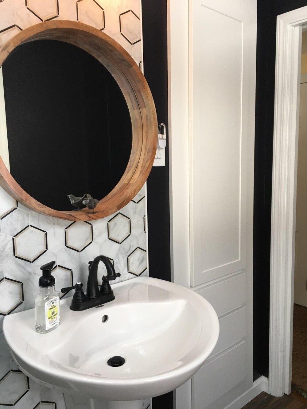 Bathroom with white pedestal sink, black faucet, round wooden mirror, and hexagon tile wall.