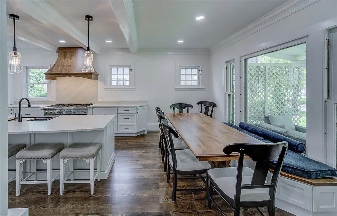 Bright kitchen with island, range, and long dining table with seating. Sunlight streams in from a large window.