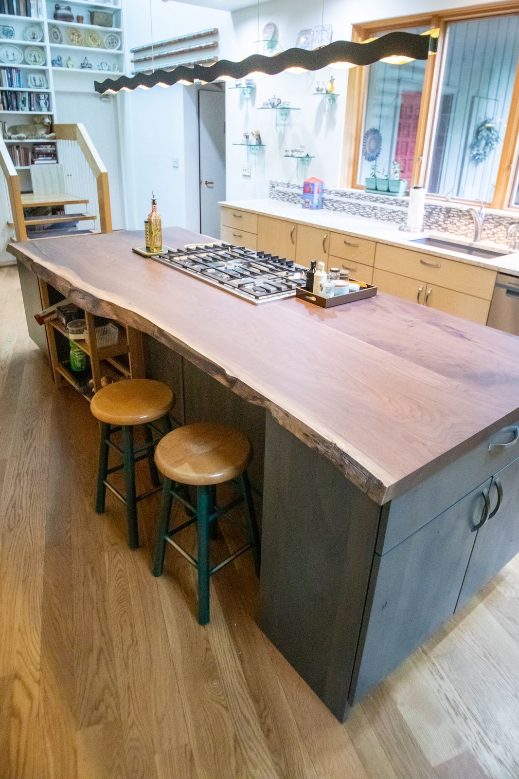 Kitchen island with live edge wood countertop, stools, and metal cabinetry. Gas cooktop, and shelving in the background.