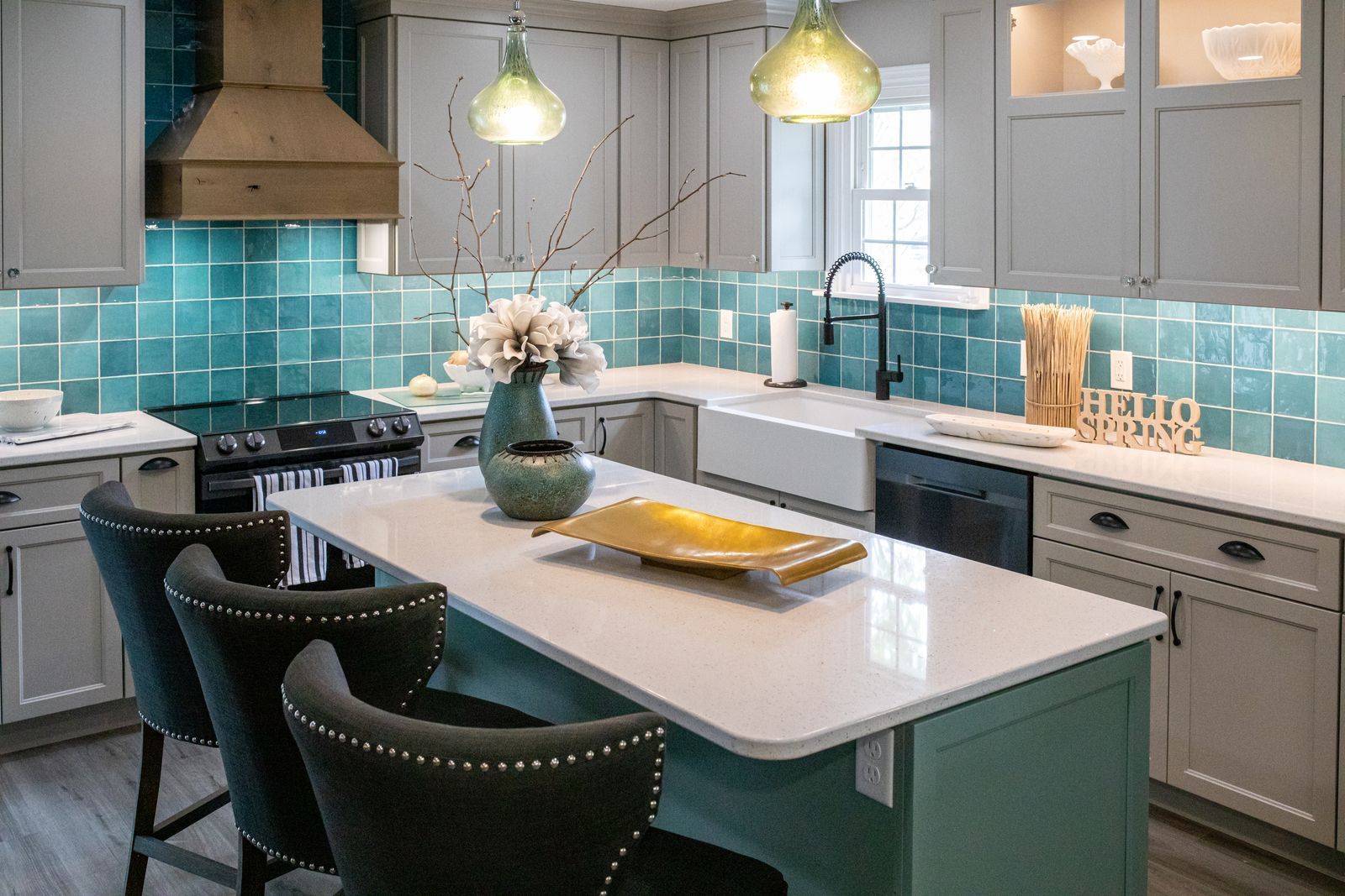Kitchen with teal backsplash, white countertops, and gray cabinets. A center island has three bar stools.