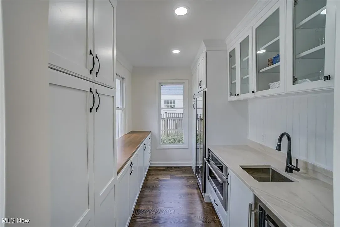 Narrow white kitchen with pantry, countertops, sink, and oven. Dark wood flooring, window at the end.