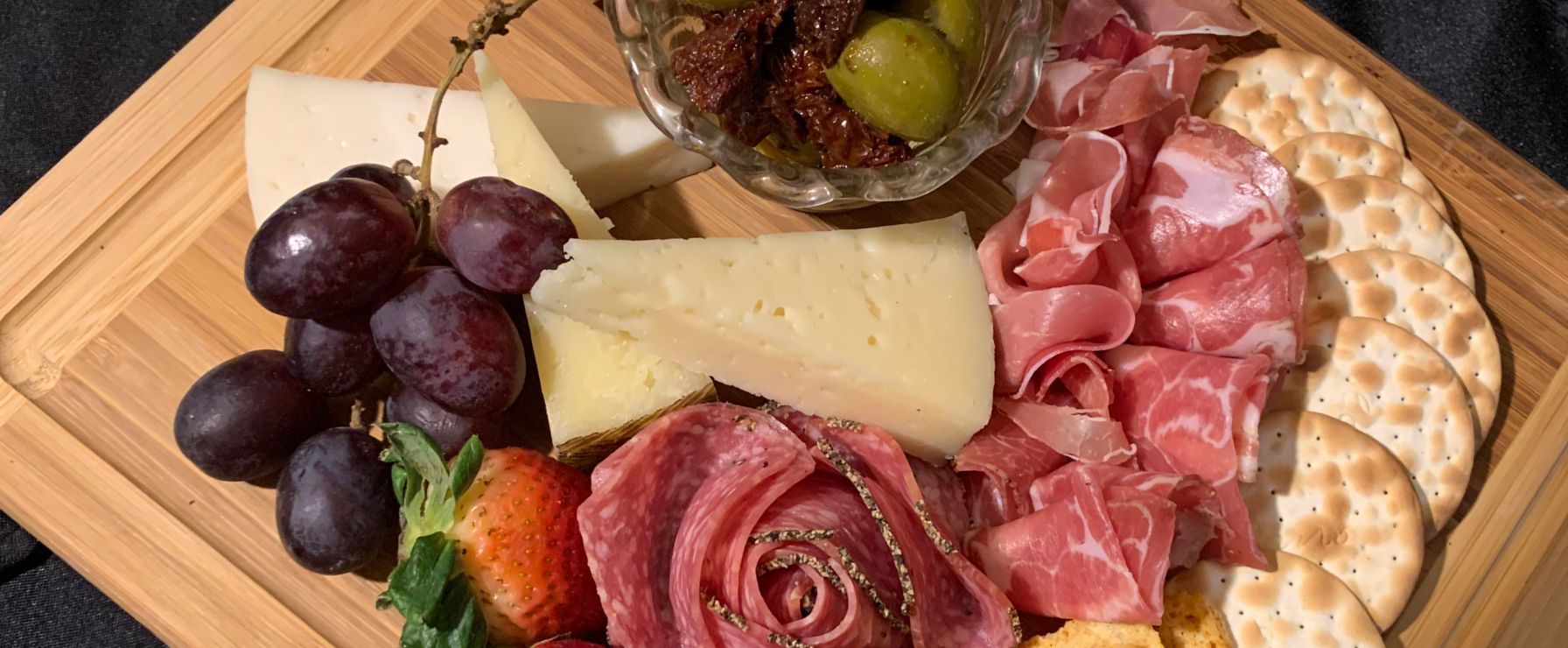 A charcuterie board featuring salami roses, prosciutto, various cheeses, grapes, olives, and round crackers.