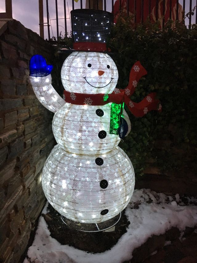 A lit-up snowman decoration with a top hat, red and green scarf, and blue mitten, standing outdoors in the snow at night.