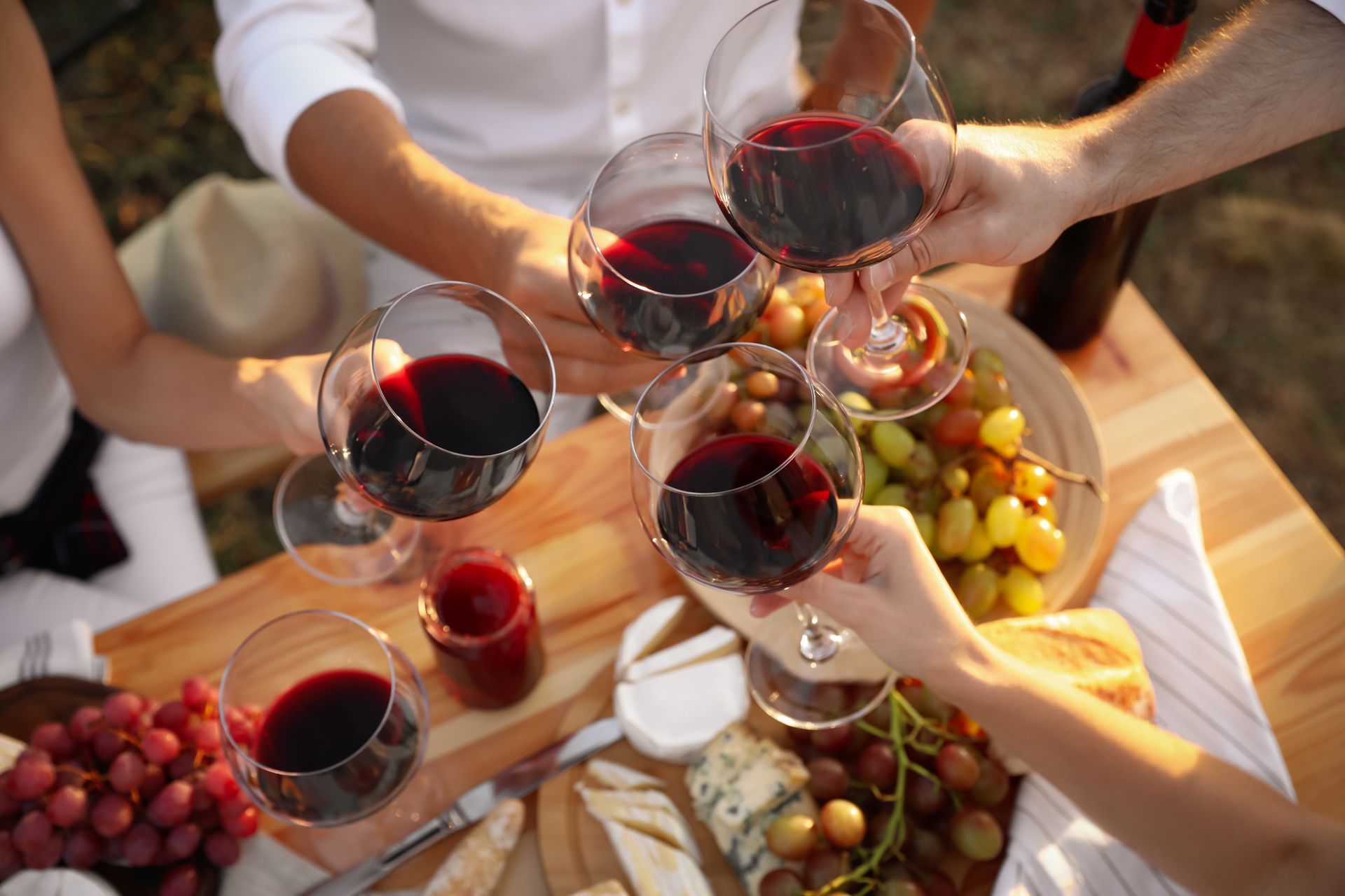 People clinking red wine glasses over a table with grapes and snacks