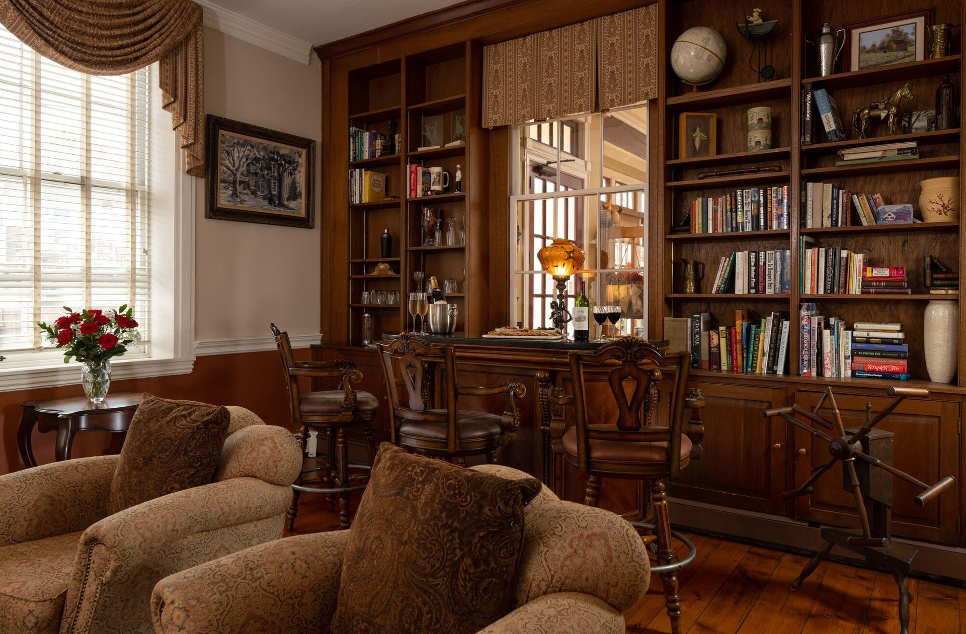 A cozy library with two plush armchairs, floor-to-ceiling wooden bookshelves, and a high-top table by a window.