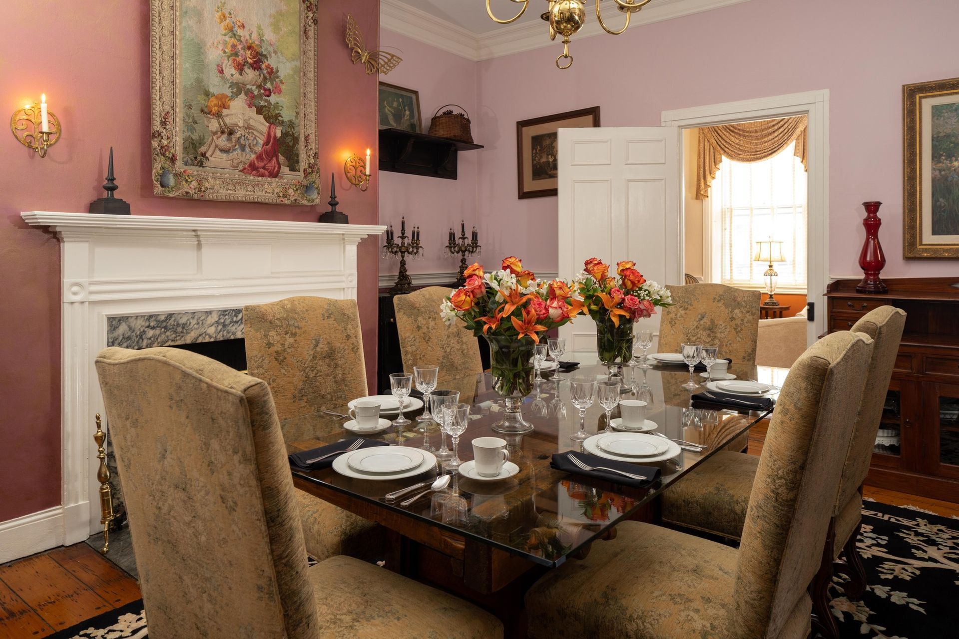 A formal dining room with a glass-topped table, tan upholstered chairs, and floral bouquets, set before a white fireplace.