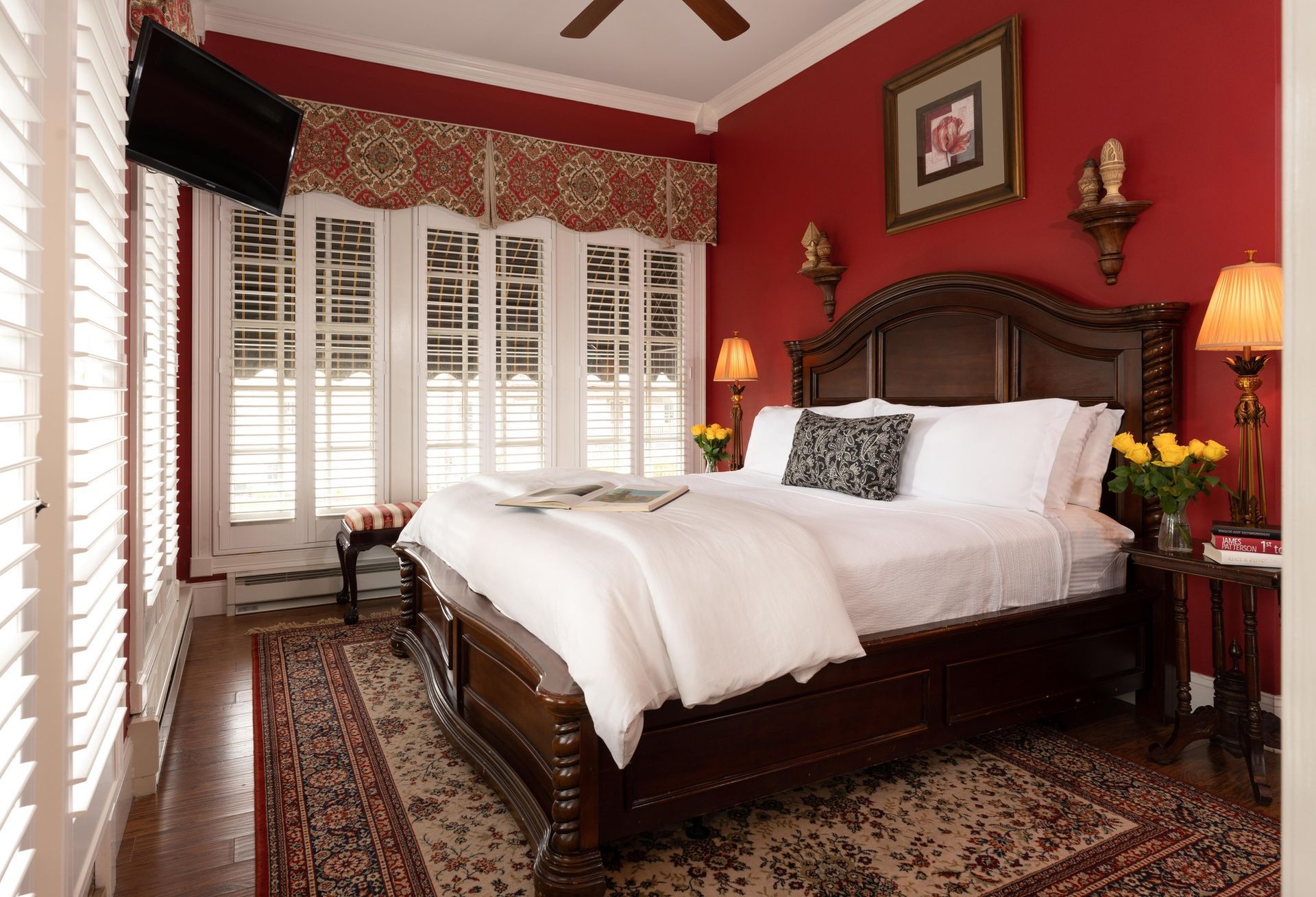 A bedroom with a large dark wood bed, crisp white bedding, red walls, shuttered windows, and a patterned area rug.