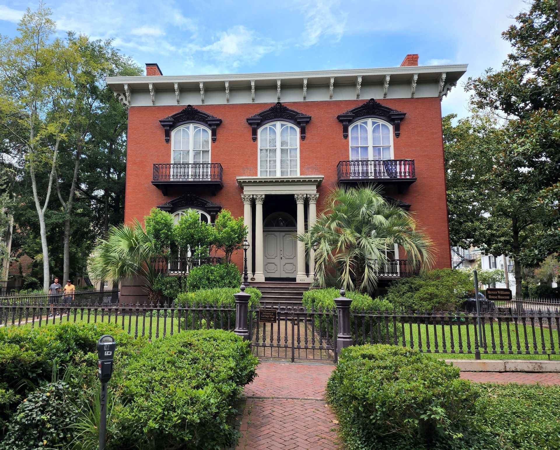 Red-brick historic house with black shutters, wrought-iron balconies, and a front garden fence.