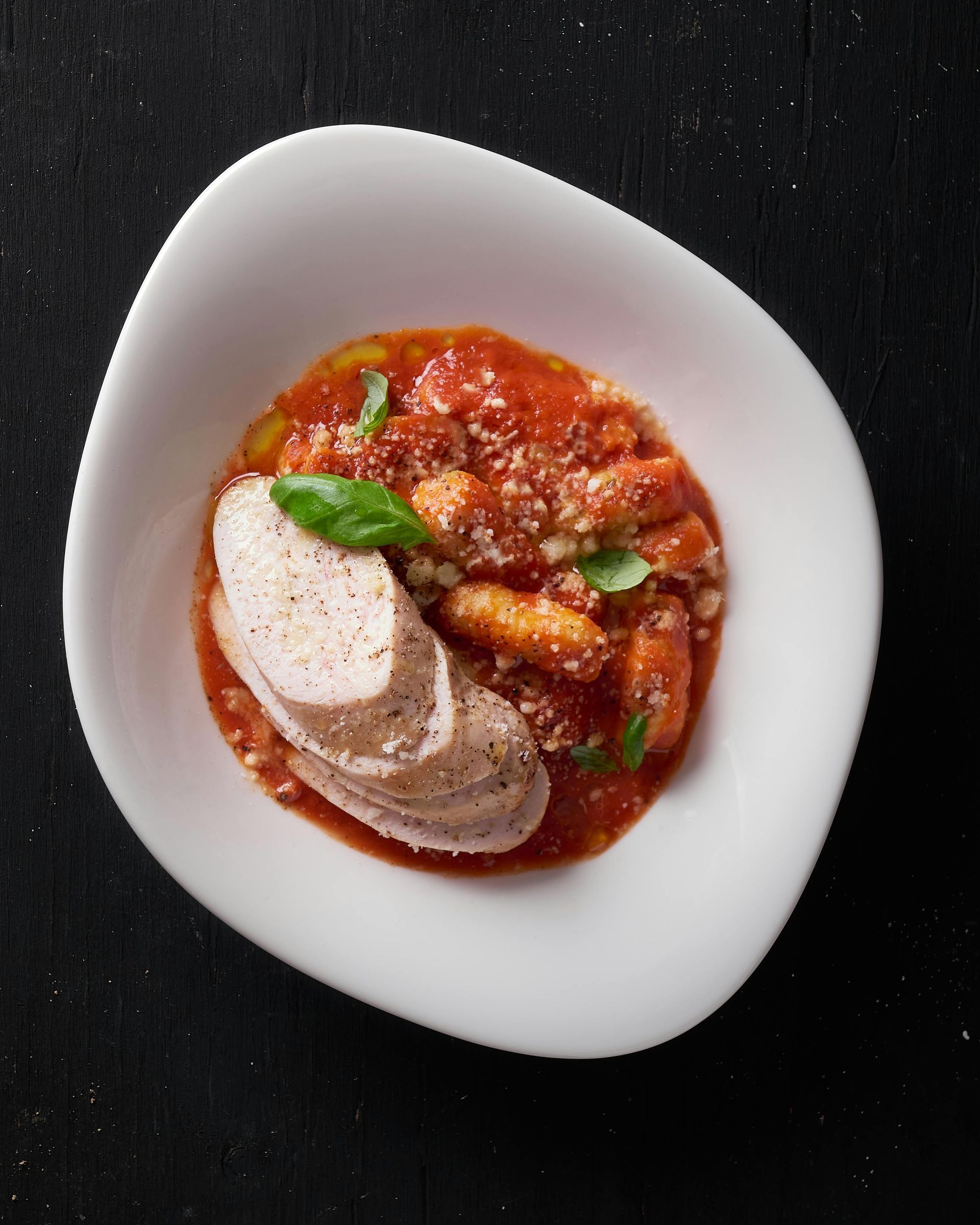 A top-down view of sliced chicken breast served over gnocchi in tomato sauce with fresh basil in a white bowl.