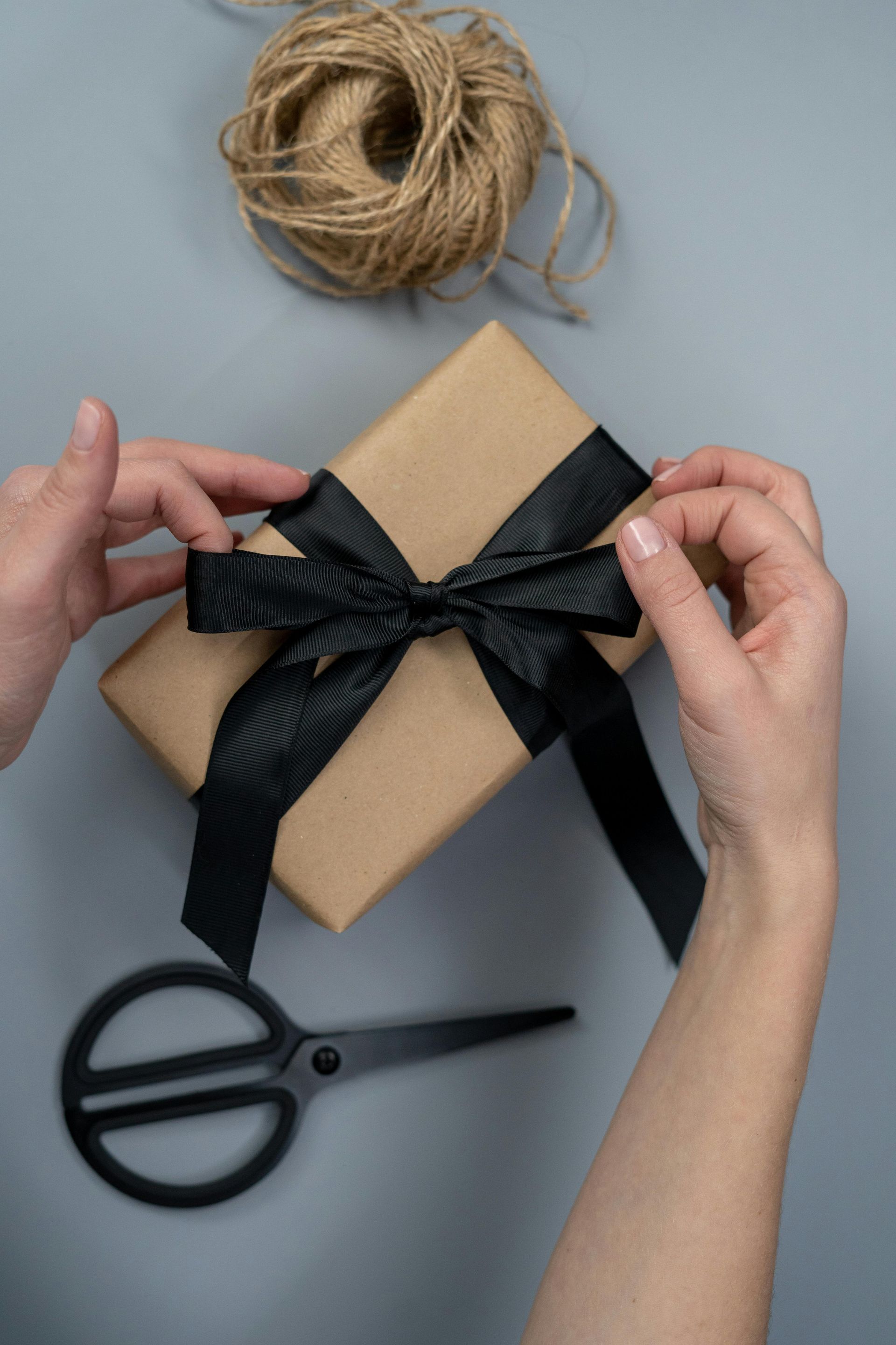 Hands adjust a black ribbon tied in a bow around a brown gift box, with scissors and twine on a grey surface.