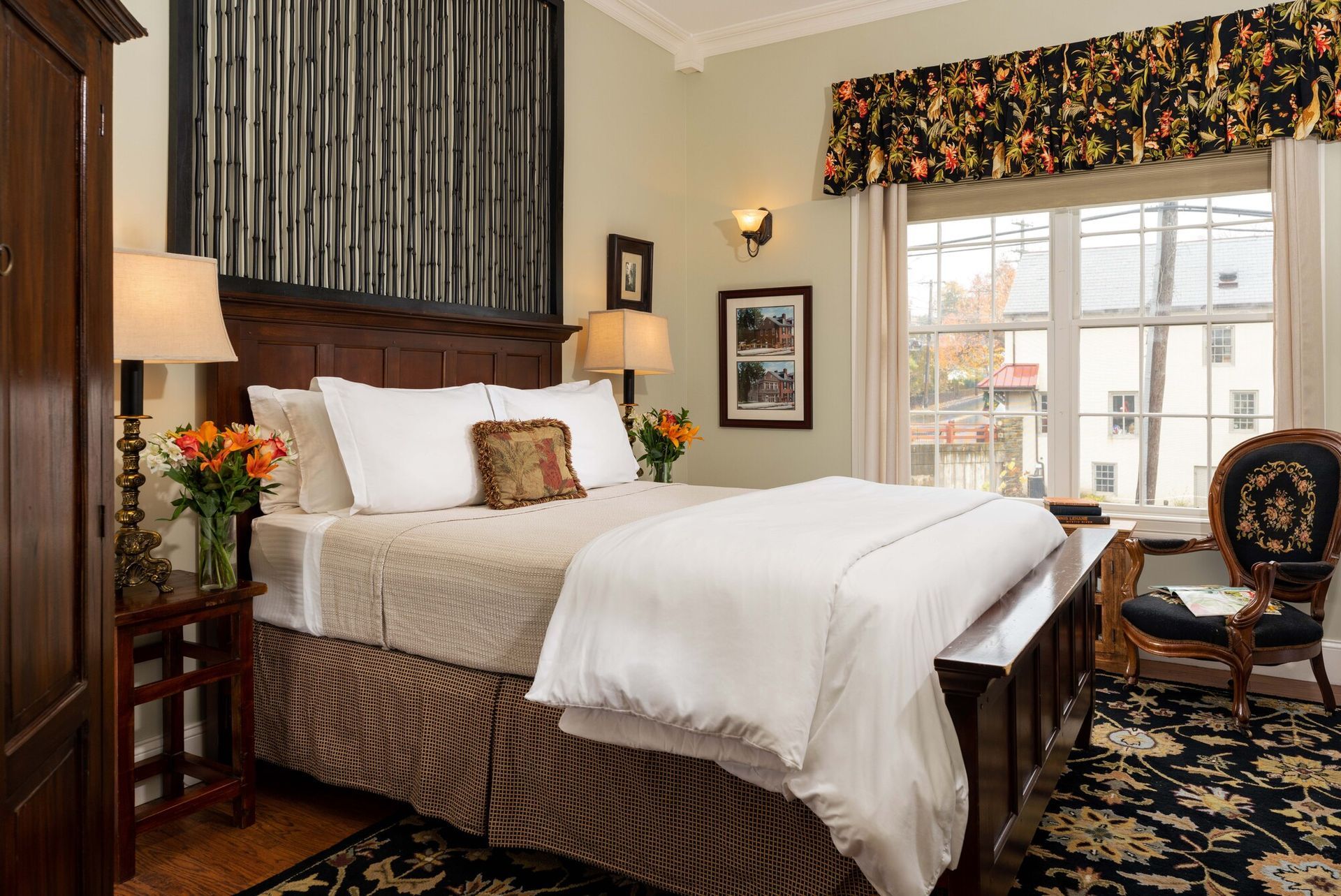 A neatly made hotel bed with white linens and a patterned skirt, flanked by wooden furniture and a classic side chair.