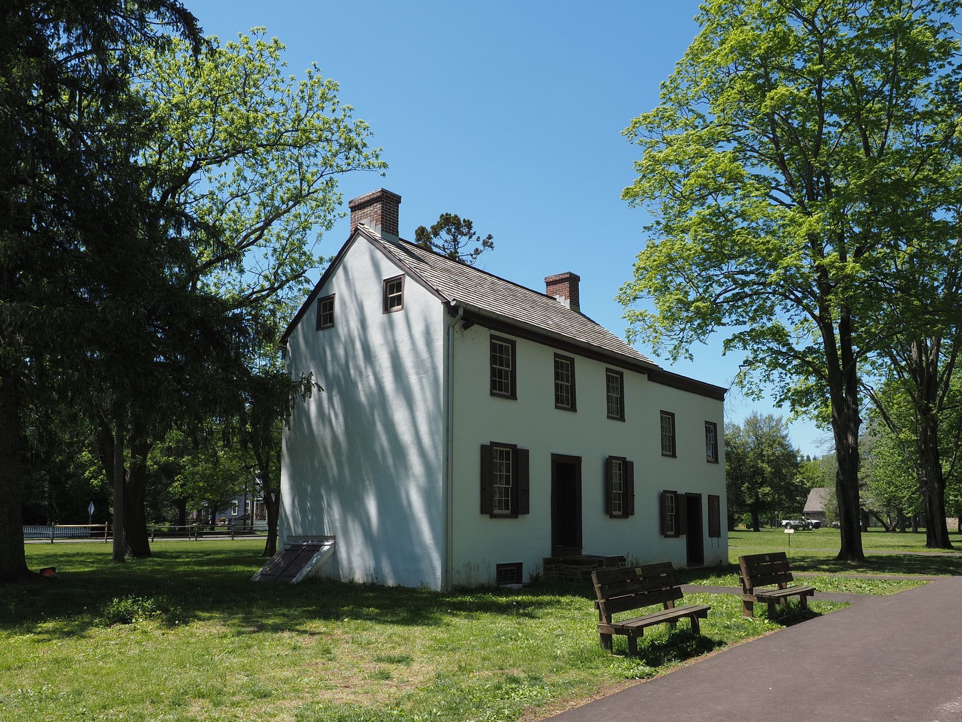White two-story house with dark shutters in a sunny park, framed by trees and a paved path.