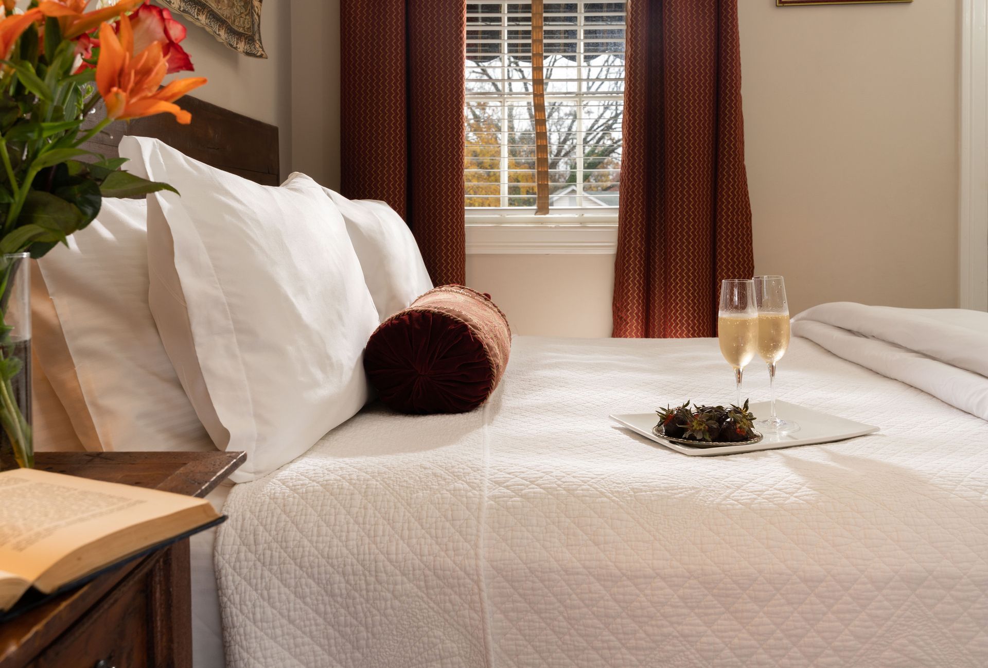 A made hotel bed with white linens, two glasses of champagne, chocolates, and a book on a bedside table with flowers.