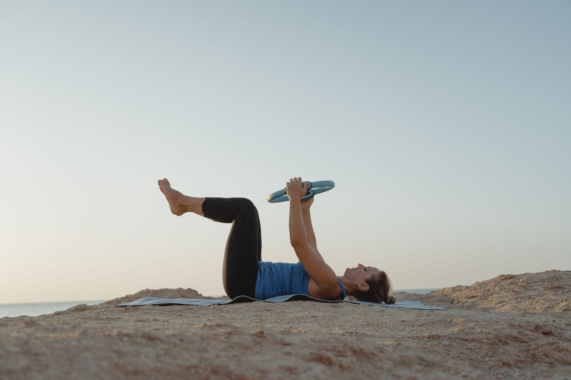 Person lying on a rock, raising bent legs and holding a turquoise dumbbell under a clear sky.