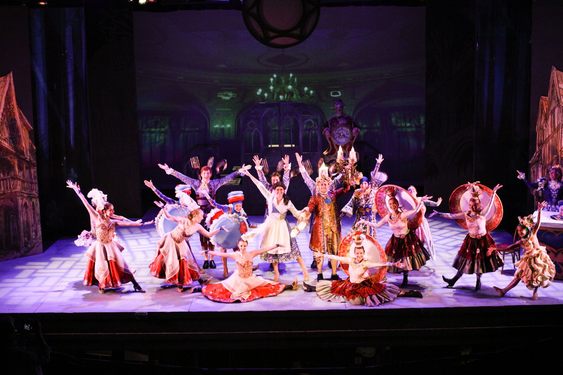 Ballet dancers in colorful costumes on stage, arms raised, with castle backdrop and side buildings.