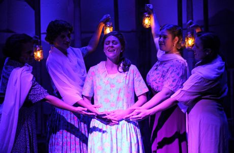 Group of people in a play, holding lanterns around a person in a floral dress, lit with blue light.