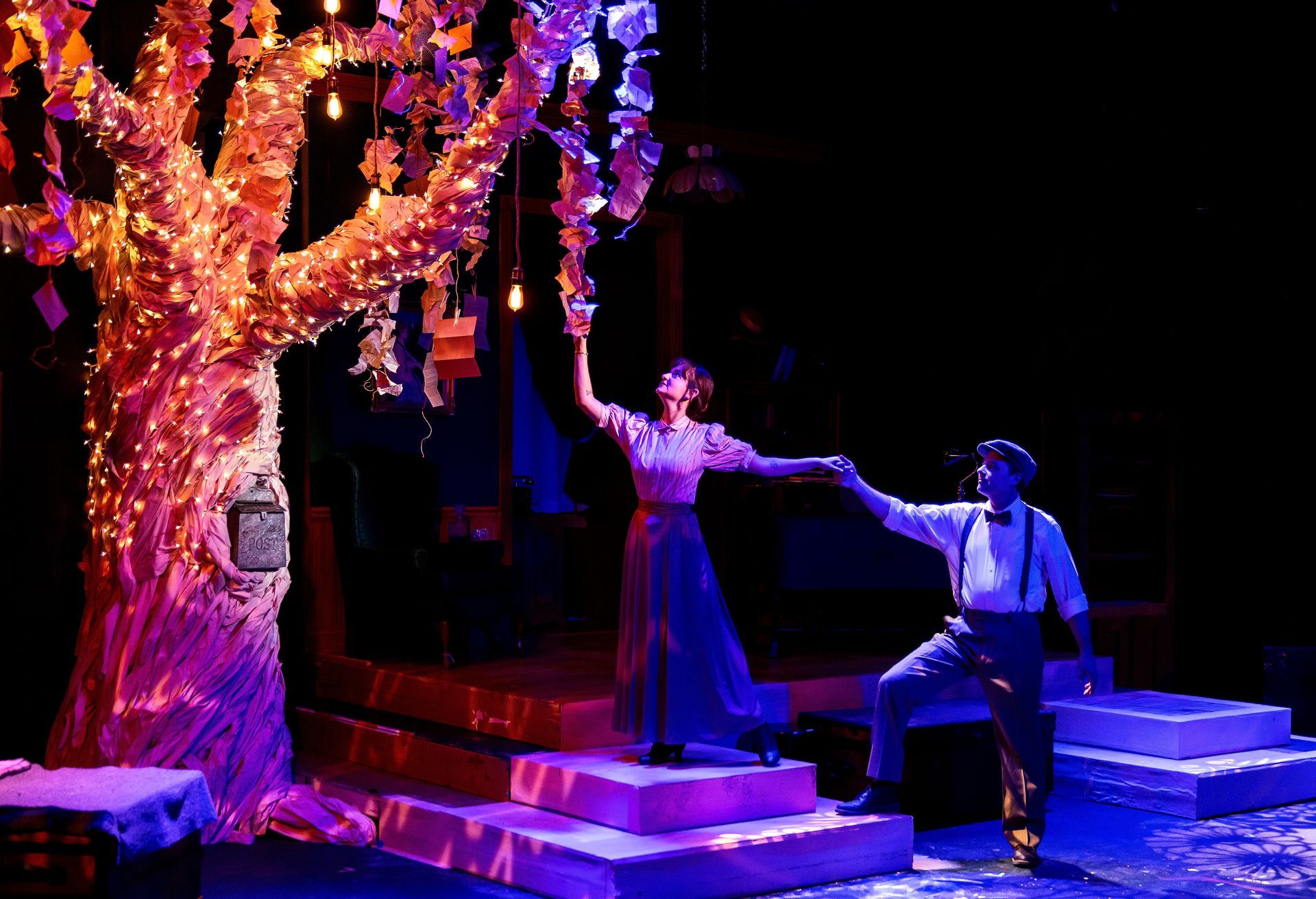 A man and woman dance on a stage, lit by a tree adorned with lights and paper.