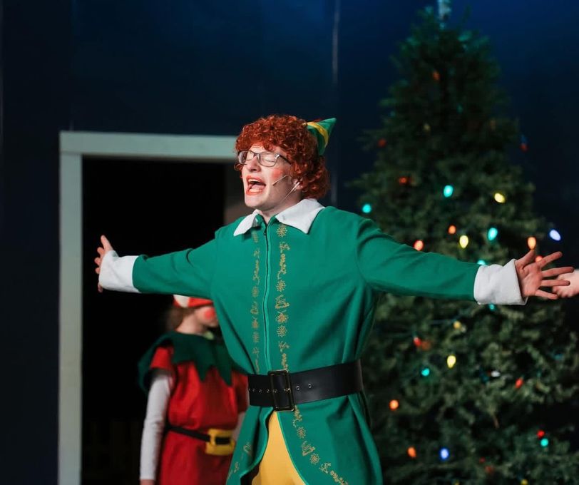 Person in an elf costume, arms outstretched, singing on a stage with a Christmas tree.