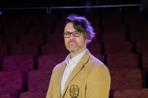 Man in tan blazer stands in theater; red seats, purple lighting.