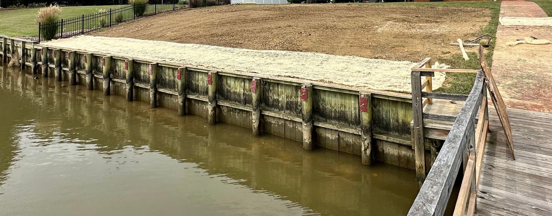A weathered wooden bulkhead along a murky waterway with dirt and gravel fill.