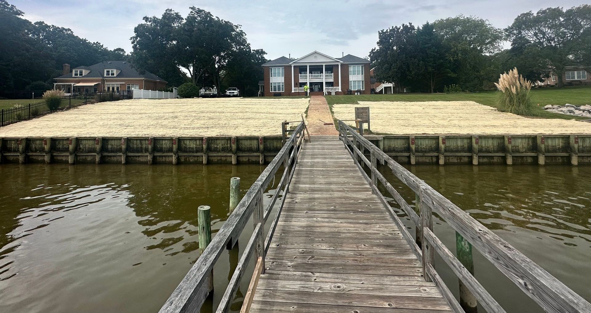 Wooden pier leads to a large brick mansion with white columns, on a sandy shoreline.