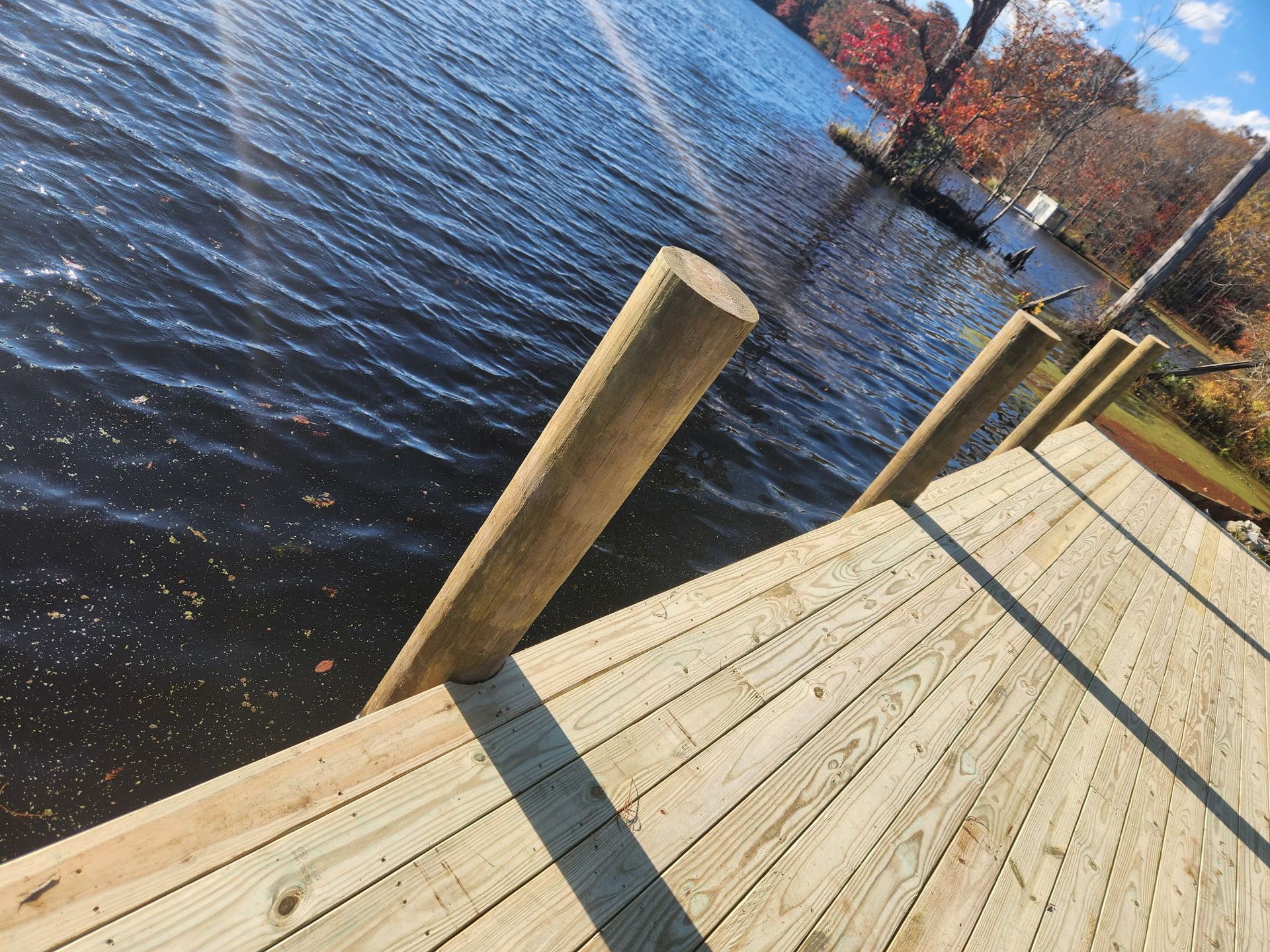 A high-angle view of a newly built wooden dock extending over dark blue lake water toward colorful autumn trees.